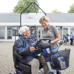Vrouw in een blauwe jas krijgt scootmobiel rijles van een fysioherapeut in een grijze polo op de parkeerplaats in Veenendaal.