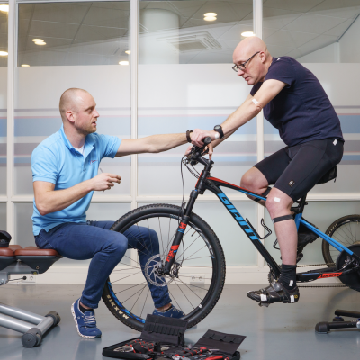 Fysiotherapeut Roel in zijn blauwe shirt helpt Evert op de fiets door middel van bikefitting om van zijn zadelpijn af te komen.
