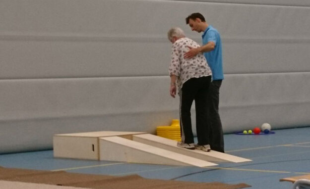 Vrouw doet aan valpreventie en wordt geholpen door een fysiotherapeut in een blauw shirt in de gymzaal.