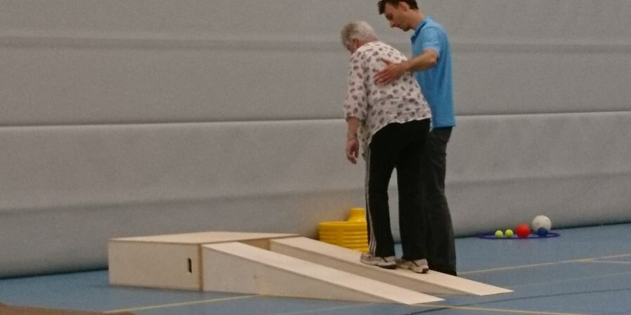 Vrouw doet aan valpreventie en wordt geholpen door een fysiotherapeut in een blauw shirt in de gymzaal.