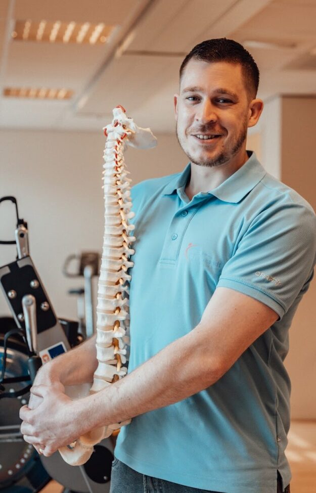 Een fysiotherapeut met een lichtblauw t-shirt aan staat in een oefenzaal. Hij houdt de wervelkolom van een skelet vast en kan zo aspecifieke rugklachten uitleggen.