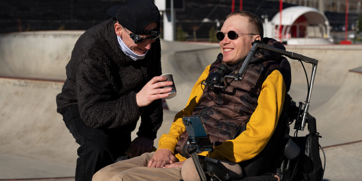 ALS Man met ALS in een bodywarmer en gele mouwen wordt geholpen door een man in een zwarte jas