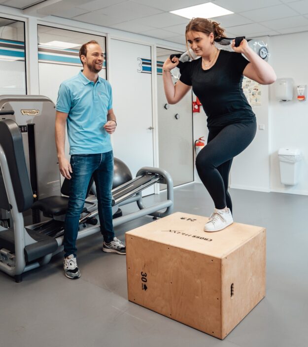 Vrouwelijke patiënt in zwarte kleding werkt weer aan de opbouw van haar spierkracht na het oplopen van een blessure met behulp van een fysiotherapeut in een blauwe polo